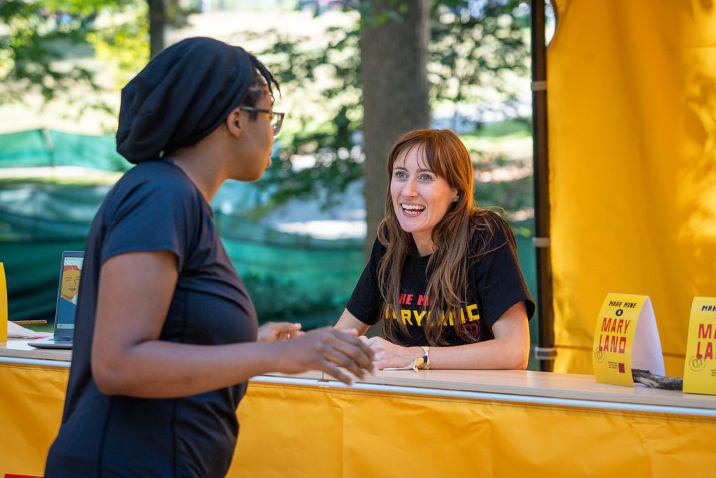 Two people talking at a yellow information table with signs that read “Make Mine Maryland.”