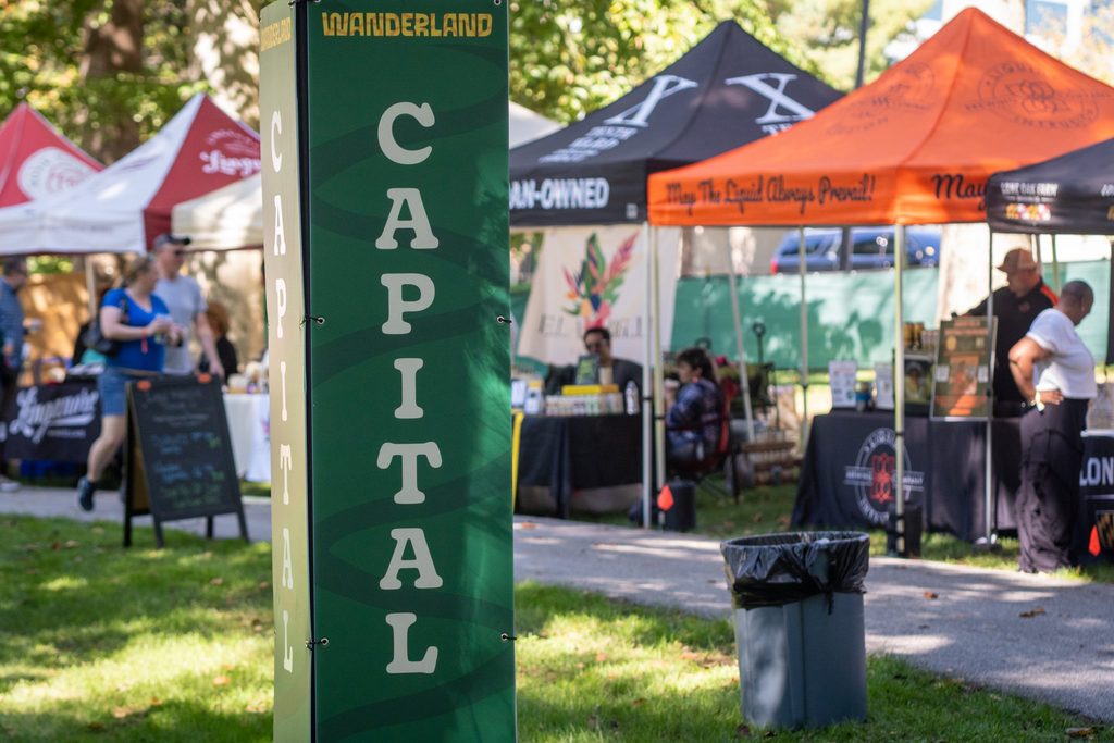 A green display pillar labeled “WANDERLAND” and “CAPITAL” standing in an outdoor market with various vendor tents in the background.
