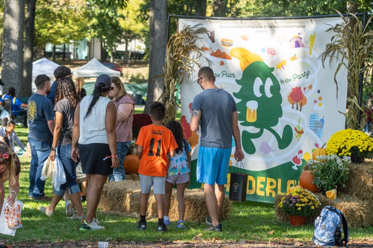 People stand in a park looking at a festive event banner featuring a cartoon pine tree holding a beer mug, surrounded by hay bales and potted mums.